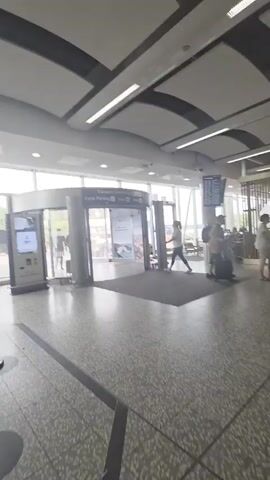 This is a video recording of an indoor environment, likely an airport terminal or a large public transit station, given the presence of a revolving door, signage, and people with luggage. The scene is well-lit with artificial lighting and natural light streaming in from large windows. The architecture features a modern design with a curved, slatted ceiling and tiled flooring.

Several individuals are visible, walking with luggage and navigating the space. Some appear to be travelers, while others may be staff or shoppers. There's a sense of movement and activity as people pass through.

Notable details include:

*   **Signage:** A sign above the revolving door indicates "Cycle Parking." Another sign prominently displays "BURGER KING." A smaller A-frame sign for "NERO" with an image of a drink is also visible.
*   **Objects:** A digital information screen is present near the entrance. A stroller is visible near the Burger King.
*   **Setting:** The environment suggests a public building with commercial services, consistent with an airport or train station. The clean, modern aesthetic points to a contemporary facility.

The time of day appears to be daytime, evidenced by the natural light. The overall atmosphere seems neutral, with no strong emotional cues discernible. The location context of Wrington, United Kingdom, further supports the interpretation of this being a travel hub.