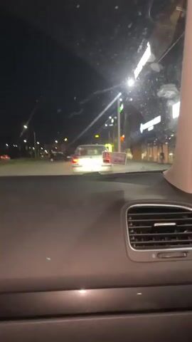 This video captures a night-time drive through Strumivka, Ukraine. The perspective is from inside a vehicle, looking forward and to the sides through the windshield and side windows. The scene appears to be a commercial area with businesses lining the street. Several cars are visible on the road, and streetlights illuminate the surroundings. 

Notable details include bright, illuminated signs on the buildings, some of which are partially obscured or out of focus. One sign appears to display the words "FOR FOOD" in large, yellow lettering. Another sign shows a blue logo with a white circular element. The car's interior is also visible, with a dashboard air vent in the foreground. The night is dark, with the artificial lights creating a sense of urban activity. The time of day is clearly night, and there are no indicators of specific weather conditions. The overall mood is that of a typical drive through a town at night.