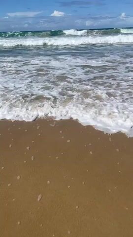 The video shows a close-up, low-angle view of gentle waves washing onto a sandy beach. The water is clear with white foam and small bubbles as it recedes. The sand appears wet and brown. In the background, larger waves are breaking with white caps, and the ocean extends to the horizon under a bright blue sky with scattered white clouds. The scene is peaceful and serene, evoking a sense of natural beauty and tranquility. There are no people or man-made objects visible in the frame.