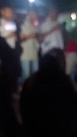 A blurry video shows a gathering of people at night. There are bright lights in the background, suggesting an outdoor event. Some people are standing and others appear to be sitting on the ground. The figures are indistinct, making it difficult to determine specific actions or interactions. The overall impression is of a casual social event or celebration.
