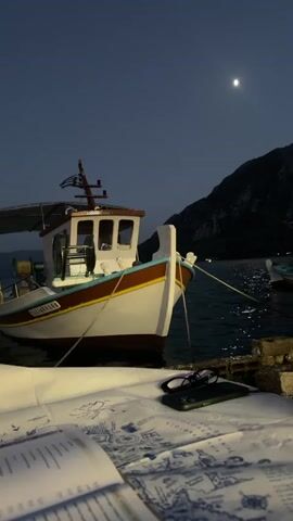 A fishing boat is docked at night in a harbor. The boat is white with a brown and blue trim. The boat is tied to a dock with ropes. In the foreground, a table is set with papers, a pair of glasses, and a mobile phone. The water is dark and reflects the moonlight. In the background, a mountain looms over the harbor. The sky is dark with a full moon.