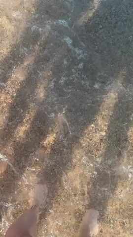 The video begins with a first-person perspective of someone walking into shallow, clear water at a beach. The camera is pointed downwards, showing the person's feet and legs as they step onto the sandy, pebbly seabed. Ripples and shadows from the sunlight play on the water's surface. The view then pans up to show the expanse of the ocean under a clear blue sky. The water is calm with gentle waves. Further into the video, a woman wearing a hat and swim attire is visible standing in the water, facing away from the camera. Other distant figures and buoys can be seen in the water. The scene depicts a typical day at the beach with a person entering the water and another person already in the sea.