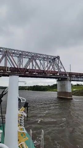 A freight train crosses a large metal truss bridge over a wide, choppy river. The camera is positioned on a boat, looking upwards towards the bridge and train. The sky is overcast and gray. The bridge is made of steel girders and supports, with railroad tracks running along the top. The train consists of many dark-colored freight cars. The river is brown and turbulent, with visible waves and whitecaps. The boat in the foreground has a white railing and a white pillar with some yellow and green equipment attached. The banks of the river are lined with green trees. The overall atmosphere is somber and industrial.