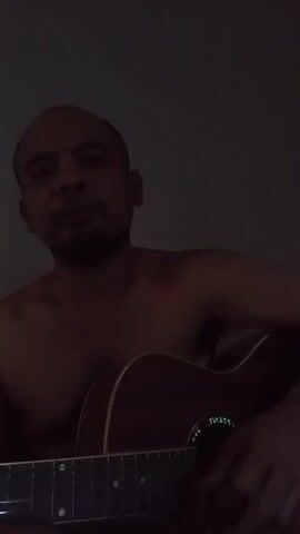 A dark, medium shot shows a bald, bare-chested man seated in front of a dark background. He is holding an acoustic guitar, with his left hand on the fretboard and his right hand near the soundhole. His mouth is open, suggesting he is singing or speaking. The lighting is dim, casting shadows on his face and body. The video is a recording of the man playing and possibly singing to the guitar.