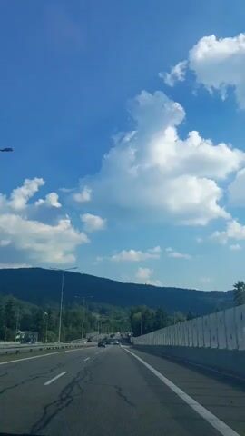 This video captures a view from a vehicle driving on a highway under a bright, partly cloudy blue sky. The road ahead is paved, with distinct white lane markings. On the left side of the road, there are streetlights and trees lining a hillside. On the right, a sound barrier, likely for a nearby residential area, stretches along the highway. In the distance, a range of tree-covered mountains forms the backdrop. The overall scene suggests a clear, possibly warm day, with the sun shining brightly. The movement of the camera indicates travel, likely on a journey through a scenic, mountainous region. The location is identified as Myślenice, Poland. There is no visible text in the video.
