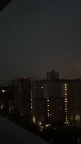 The video shows a nighttime cityscape with apartment buildings. The sky is dark, with occasional flashes of lightning and a faint purple hue in the clouds at 00:02. Lights are visible in the windows of the buildings, and streetlights illuminate the street below. In the distance, a tall industrial smokestack emits light. The video appears to be taken from a balcony or elevated vantage point.