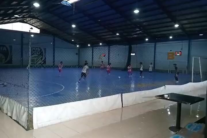 The video shows an indoor futsal court with a blue playing surface and a dark blue roof supported by metal beams. Bright fluorescent lights illuminate the court. A net surrounds the playing area, and a white padded barrier runs along the bottom edge of the net. A black table and chairs are visible in the foreground to the right. On the court, several young boys, dressed in athletic wear, are playing a futsal match. Some are wearing red and white striped jerseys, while others are in white or gray. The players are in motion, dribbling, passing, and shooting the ball. There are goals at either end of the court, and a scoreboard is visible on the far wall displaying red digital numbers. Logos, including a "FIFA" logo, are present on the walls and banners. The overall atmosphere is one of an active youth sports event.