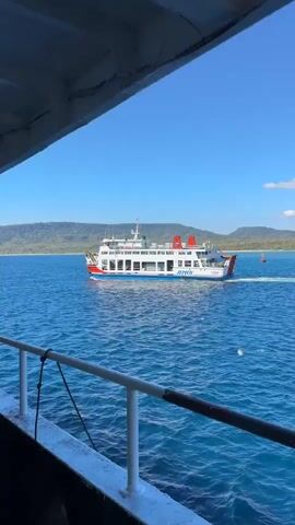 The video shows a view from a ship deck looking out at a body of water with a ferry boat moving in the distance. The water is blue and appears choppy. In the foreground, there is a railing with a rope attached. The sky is clear and blue with some white clouds. The ferry boat is white with red and blue accents and has the word "ARTIN" visible on its side. The ferry is moving from left to right in the frame. In the background, there is a distant shoreline with green, hilly land. The video then cuts to a close-up of a man wearing a black baseball cap and a grey tank top. He is looking at the camera and giving a thumbs-up gesture. He has long fingernails. The background behind him is the blue sea and sky. The man is smiling and appears to be enjoying himself.