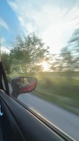 The video shows a view from inside a car as it travels down a road. The focus is on the outside scenery, visible through the car window and reflected in the side mirror. The scene depicts a rural landscape with trees and fields alongside the road. The sun is setting or rising, creating a bright glow on the horizon and casting light across the sky with scattered clouds. The car's interior, including the door panel and side mirror, are visible in the foreground.