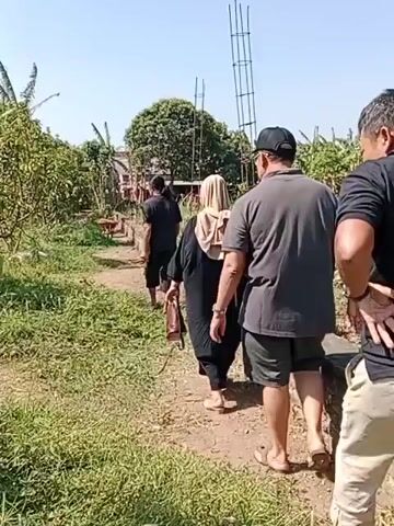 A group of four people walk along a dirt path in an outdoor environment. The path is lined with green vegetation, including trees and grass. In the background, there are buildings and a clear blue sky. The people appear to be walking away from the camera, in single file. The person in the foreground on the right is a male wearing a black shirt and light-colored pants. Next to him, to his left, is another male wearing a grey shirt and shorts. Behind them is a female wearing a headscarf and a dark outfit, carrying a brown bag. In front of her, further down the path, is a male wearing a black t-shirt and shorts. A tall, unfinished structure made of rebar is visible on the right side of the frame. The overall scene suggests a rural or semi-rural setting.