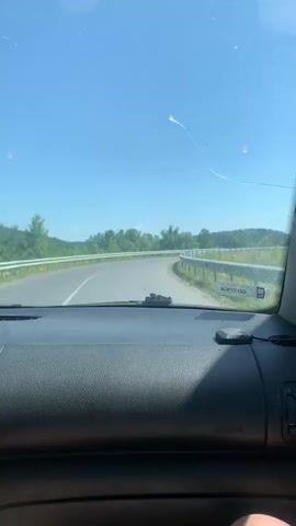 The video shows a view from inside a car looking out the windshield on a sunny day. The car is driving on a paved road with a white guardrail on the right side. There are trees and hills in the background. The sky is clear and blue. There are some smudges and a few small scratches on the windshield.