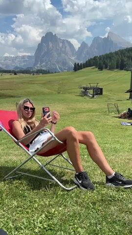 A young woman with blonde hair, wearing sunglasses, a black tank top, and white shorts, is sitting in a red lounge chair outdoors. She is holding a smartphone in her left hand and appears to be taking a selfie. The chair is placed on a grassy field. In the background, there are rolling green hills, trees, mountains, and a ski lift. The sky is blue with scattered white clouds. The lighting suggests it is daytime and sunny.