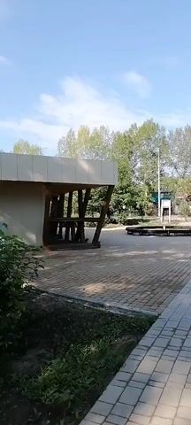 The video shows a panning shot of an outdoor park area on a sunny day. In the foreground, there is a paved walkway made of interlocking rectangular stones. To the left, there is a patch of green grass and bushes.

In the middle ground, the walkway continues into a wider open space with more paving. To the left of the walkway, there is a modern-looking building with a slanted roof supported by diagonal wooden beams. Beyond this structure, trees and foliage are visible. Further into the park, a circular metal structure, possibly part of an art installation or playground equipment, is present. There is also a tall pole with a flag or sign attached. Benches are scattered throughout the area.

To the right of the walkway, another paved area leads further into the park. A tall, slender wooden sculpture or pillar stands prominently. More trees and greenery line the sides of the path. In the distance, more park structures and trees can be seen under a clear blue sky with scattered clouds. The overall impression is of a well-maintained public park or recreational area. No people are visible in the video.