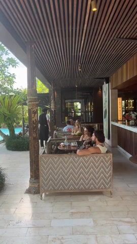 The video shows a tropical resort.  It starts with a shot of people sitting and drinking at an outdoor restaurant, focusing on a couple at a table. A waiter walks by. The camera pans to the left to show the edge of a pool.  It shows the clear, blue water of the pool surrounded by tropical plants and trees.  People are swimming and relaxing in the pool. The sky is blue with a few fluffy white clouds.