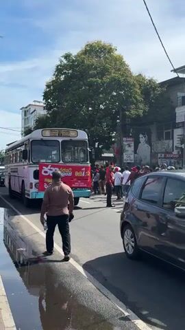 The image captures a street scene in Delkanda, Sri Lanka, on a bright, sunny day. A white public transport bus with green stripes, typical of Sri Lankan Transport Board (SLTB) vehicles, dominates the left side of the frame. The bus displays "138" as its route number, with "PILIYANDALA" and "KOTTAWA" (in both Sinhala and English) also visible, indicating its destination.

A large red banner with white Sinhala text is prominently displayed across the front of the bus. The text reads "ජන ව්‍යාපාරය" (Jana Vyaparaya - People's Movement/Campaign) and "මැයි 1" (May 1st), strongly suggesting a May Day (Labour Day) rally or political demonstration.

A crowd of people, many wearing red shirts, are gathered in the street around the bus, some appearing to walk alongside it. This collective attire further supports the notion of an organized event. One man in a brown shirt and black pants walks parallel to the bus on the left, holding a water bottle. Another individual in a red uniform, possibly a bus conductor or event organizer, stands near the bus's front door.

The asphalt road shows wet patches, possibly from recent rain or overflowing water. Buildings with shops, including one displaying "SERVO OIL," line the background. A large green tree provides shade, and a mural or poster of a person is visible on one of the buildings. A dark-colored car is partially visible on the right side of the frame. The scene portrays an active urban environment centered around a public gathering.