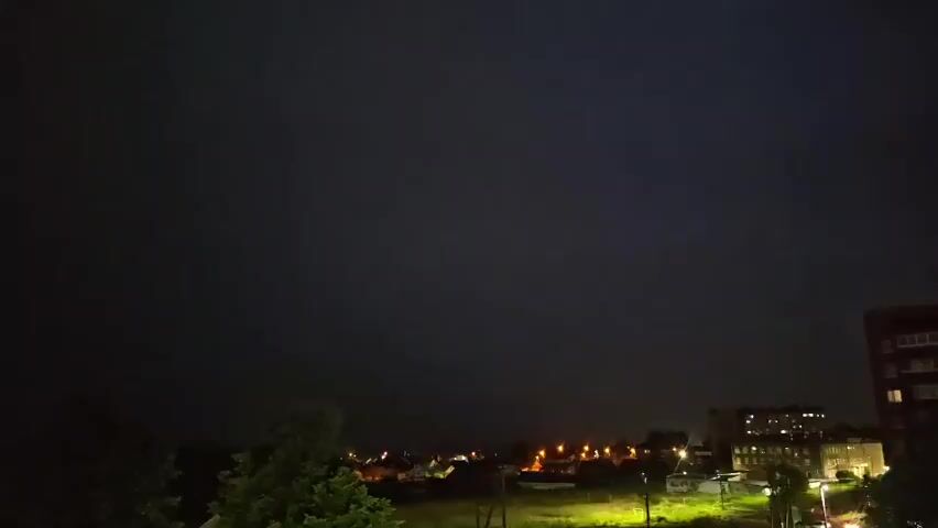 The video captures a dark, stormy night scene from a high vantage point. The sky is predominantly black, with faint hints of distant lightning illuminating the scene intermittently. Below the dark sky, a small town or village is visible. Streetlights cast a warm glow on the houses and a grassy field in the foreground. A few trees are silhouetted against the night sky on the left side of the frame, and a multi-story apartment building is visible on the right. The overall atmosphere is ominous, with the occasional flashes of lightning suggesting an ongoing thunderstorm.