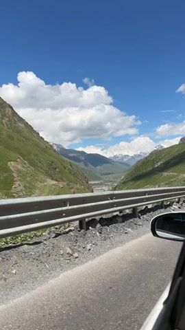 This is a POV video from a car driving on a road through a mountainous valley. The road is paved and has a metal guardrail. The valley is green with grassy hillsides and a river or stream visible in the distance. Snow-capped mountains are visible in the background under a blue sky with white clouds. The sun is shining.