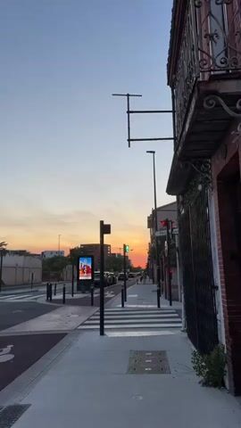 The video presents a street scene during sunset. The sky is a mix of orange, pink, and blue hues. On the right side of the frame is a building with a metal gate, and an overhang decorated with greenery. On the left is a street with cars parked along it, and a tramline in the foreground. A crosswalk spans the width of the street. Traffic lights are visible, some showing green, others red.  Street lamps line the road, and a digital billboard is present near the crosswalk.  The scene is generally quiet, captured from a static viewpoint on the sidewalk.