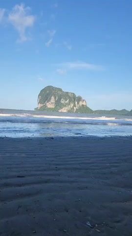 This is a vertical, outdoor, full-shot video of a beach with a calm sea and islands in the distance. The sky is blue with scattered clouds. The sand on the beach has ripples from the receding tide, and there are some shells and debris scattered across it. In the middle ground, there are two dogs on the beach, one black and one brown. One dog is standing and looking toward the water, while the other is walking away from the camera. The video appears to have been taken during the daytime, likely in the morning or late afternoon given the lighting.