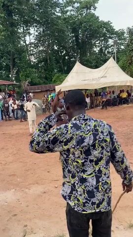 The video depicts an outdoor communal gathering in Achara, Nigeria, under what appears to be an overcast or partly cloudy daytime sky. The setting is a unpaved area with reddish-brown dirt ground, surrounded by lush green trees and vegetation, indicative of a rural or semi-rural environment.

In the foreground, a man wearing a black, yellow, and white patterned long-sleeved shirt is seen from behind, moving rhythmically. He holds a stick or whip in his right hand, suggesting he is dancing or performing as part of the event. Another man, wearing a blue and white striped shirt, black pants, and slides, with a light green cloth draped over his shoulder, walks towards the left, gesturing with a raised hand and smiling, seemingly engaging with the crowd or participating in the performance.

A significant crowd of people, comprising adults and children, is gathered in the background, observing the activities. Some spectators are seated under a large white tent, while others stand around. The tent has partially visible, inverted text on it that reads "OBIMRA AGE GRADE". A tall pole, possibly a light or speaker stand, is visible near the tent. The overall mood appears celebratory and engaging.