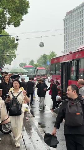 The video shows a street scene in China on a rainy day. Several red buses are parked along the side of the road, and people are disembarking and boarding. Pedestrians are walking along the sidewalk, some carrying umbrellas or bags. The street is wet from the rain. A Hampton by Hilton hotel can be seen in the background. The overall impression is one of a busy urban scene with people going about their daily activities in inclement weather.