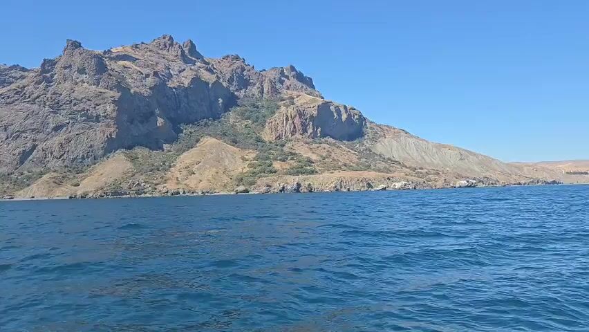 The video shows a view from a boat on the water, looking towards a rocky mountain range under a clear blue sky. The mountains are rugged and appear to be barren, with some scattered vegetation on their slopes. The sea is calm with small waves. The video pans slightly to the left, revealing more of the coastline and the extensive mountain range. The light suggests it is daytime, likely mid-morning or afternoon. There are no people or man-made objects visible in the shot, other than the implied presence of the boat from which the video is being recorded.