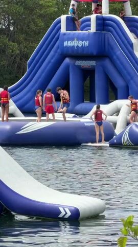 A group of young people in swimsuits and life vests are on and around a large inflatable water park structure in a body of water. The structure is blue and white and has slides and climbing areas. Some of the individuals are standing on platforms, while others appear to be interacting with each other. One person is standing on a white inflatable with blue trim that appears to be a slide. Another person is standing on a large blue inflatable with "MAMMOTH AQUAPARK" written on it. The water is calm and appears to be a lake or pond. Trees are visible in the background.