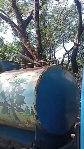 A long, horizontal metal tank, painted with a mural of trees and flowers, is being filled with water. The tank appears to be mounted on a vehicle, visible at the bottom of the frame. Water is pouring from a pipe into an opening on the top of the tank. The scene is set outdoors, with trees and foliage visible in the background. The tank is rusted in places, and the paint is faded and chipped.