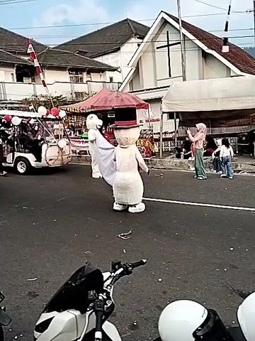 A parade is taking place on a street. In the foreground, a motorcycle is partially visible. In the middle ground, two individuals in mascot costumes, resembling a snowman and a rabbit, are walking. Behind them, a black pickup truck decorated with red and white streamers and flags is moving, with people riding in the back. To the left, a white motorized rickshaw, also decorated, is part of the procession. Several people are watching the parade from the side of the road, some are holding cell phones and taking pictures. In the background, there are buildings, including one with a cross on top, likely a church, and trees. The sky is overcast. The overall atmosphere suggests a festive event.