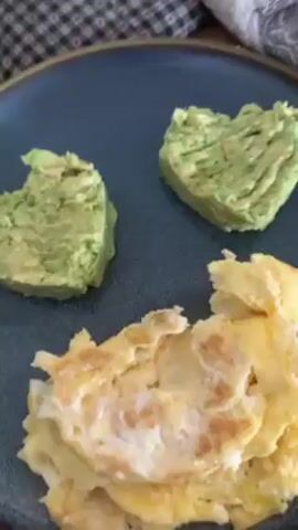 A man in a red long-sleeved shirt is seen spreading mashed avocado from two heart-shaped mounds onto a pile of scrambled eggs on a dark blue plate. He uses a small knife with a purple handle to spread the avocado. The plate sits on a light brown table. The man wears glasses and has short grey hair.