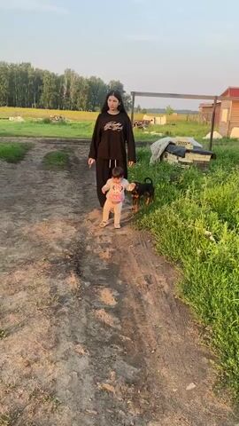 A young woman in a black sweatshirt and black pants stands on a dirt path. She is looking down at a baby in a light-colored outfit who is walking ahead of her, holding out their arms for balance. A small brown and black dog walks alongside the baby. In the background, there is a field with trees, a wooden fence, and a building. To the left, a red ATV with several people on it is driving away. The sun is low in the sky, casting a warm light on the scene.