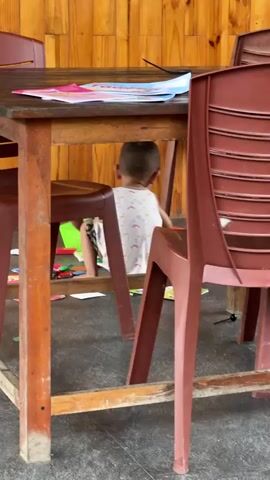In an indoor or covered outdoor setting in Du Già Commune, Vietnam, a young child, seen from the back, is sitting on the floor under a wooden table. The child is wearing a white tank top with subtle patterns and shorts.

On the wooden table, there are some papers or a book. Several brown plastic chairs are positioned around the table. Underneath the table, a green plastic basket holds numerous colorful small objects or toys, which are also scattered on the dark-colored floor.

The child is actively playing, reaching into the green basket, grabbing a handful of the colorful toys, and then letting them drop onto the floor, creating a distinct clattering sound. The background features a wall made of light-colored wooden panels. The scene suggests a casual and playful moment.