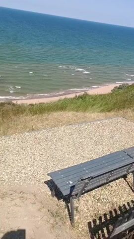 This media file depicts a scenic view of the sea from a slightly elevated vantage point. In the foreground, a grey park bench is visible, positioned on a gravel-covered area. Beyond the bench, a grassy slope leads down to a sandy beach where gentle waves are breaking. The sea itself is a vibrant turquoise, stretching out to meet a clear, pale blue sky. The overall impression is one of a peaceful, sunny day at the coast. The time of day appears to be midday or early afternoon, given the bright light and clear skies. There is no visible text in the media file. The setting suggests a public space, possibly a park or viewpoint overlooking the sea in Kuchugury, Russia. The serene atmosphere evokes feelings of tranquility and relaxation.