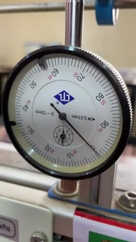 The video shows a dial indicator up close. The dial face is white with black and red markings and a blue logo. A long black needle indicates a value on the outer dial, while a smaller needle indicates a value on a smaller dial within the larger one. The dial indicator is mounted on a metal stand with various other components and markings visible in the background. The overall setting appears to be a workshop or laboratory.