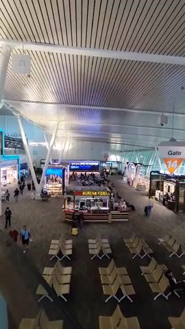 The video shows a wide shot of an airport departure lounge. The lounge has a modern design with a high, slatted ceiling and large windows overlooking an outdoor area. There are rows of empty beige chairs facing away from the camera, and some people are seated in the chairs further down the lounge. A food stall labeled "BURGER & GRILL" is visible in the center of the frame, with a few customers standing in front of it. Further down the lounge, there are several shops and kiosks, and people are walking around. A sign indicating "Gate 14" hangs prominently in the air. The overall atmosphere appears calm and ordinary for an airport environment.