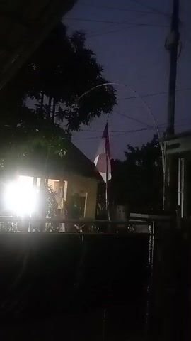 The video begins in darkness, with a bright light illuminating the left side of the frame, likely from a house. A flagpole with a flag, partially obscured, stands in the middle ground. The flag appears to be red and white, possibly the Indonesian flag. A string of lights curves over the flagpole. As the video progresses, the lights change color, cycling through white and red. Trees and parts of buildings are visible in the background, all silhouetted against a dark, twilight sky. The overall impression is of a residential outdoor scene at dusk or night.