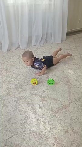 A baby is lying on its stomach on a patterned carpet. The baby is wearing a dark t-shirt with yellow trim and black shorts. Two small, colorful toys are placed on the carpet in front of the baby. One toy is yellow with a winking smiley face, and the other is green with a smiling smiley face. The baby appears to be attempting to reach for the toys, pushing up with its arms. The background shows a white curtain and a beige wall. The lighting suggests an indoor setting during the daytime.