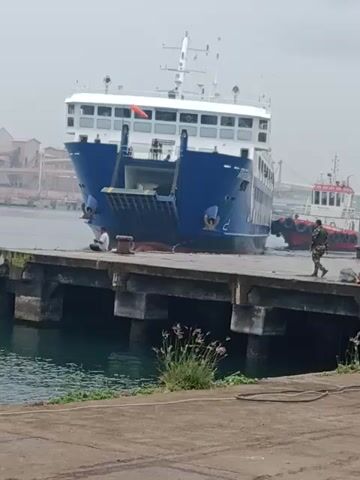 A vertically oriented, medium shot captures a blue and white ferry docked at a concrete pier.  The pier is constructed with blocky concrete supports extending into the water.  A small patch of greenery grows along the edge of the pier closest to the camera.  Several figures are visible on the pier, appearing to be workers, but their actions are difficult to discern. Another smaller, red and white boat is moored behind the ferry. In the background to the left, industrial structures are partially visible. The sky is overcast, creating a flat lighting condition.