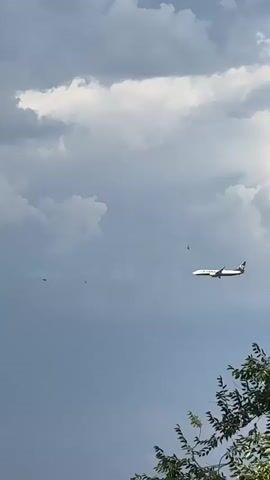 A passenger airplane is shown in flight against a cloudy sky. It appears to be a twin-engine jet with white fuselage and a dark tail fin. The plane is in the middle distance, and the camera is looking up at it from below. In the foreground, there are trees with green leaves. The sky is mostly overcast with some darker storm clouds in the background. There are also a few distant buildings visible at the bottom of the frame, suggesting an urban or suburban setting. The plane is moving from left to right across the frame. There are no people visible in the video.