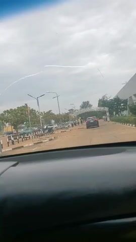 The video shows a car driving through a parking lot toward a gated entrance. The sky is overcast, and there are street lights and trees. Cars, motorcycles, and people are visible in the background. A box of tissues and a decorative figurine are on the dashboard.