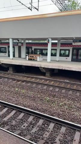 The video shows a train station platform. The platform is covered by a white, flat roof supported by white pillars. A white train with a decorative red pattern along the top is stopped at the platform. The train doors are marked with green lines. A person wearing dark clothing is walking on the platform. There is a wooden bench on the platform. Below the platform are railway tracks covered in gravel and weeds. Overhead power lines and support structures are visible above the platform roof. The sky is overcast.