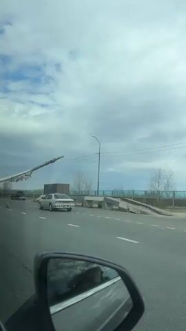 From the interior of a vehicle, the video shows a very large wind turbine blade being transported on a multi-wheeled trailer. Traffic cones are placed around the trailer and a few other vehicles are visible on the road. The sky is cloudy. The side mirror of the car recording the video is visible on the left.
