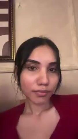 A woman with dark hair pulled back and parted in the middle, wearing a red top, is looking at the camera. She purses her lips slightly and then opens her eyes. In the background, a framed picture hangs on the wall. The wall appears to be off-white or light beige.