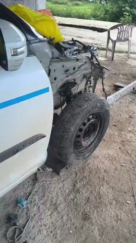 The video shows the front of a white car that has been heavily damaged. The hood is open, revealing the engine and various components. The front bumper and fender appear to be missing, and the chassis is exposed. A tire is still attached to the front wheel assembly, which is resting on a metal support. The scene appears to be outdoors, with dirt and debris on the ground. There are no people visible in the video. The overall impression is that of a car that has been in a significant accident and is in the process of being dismantled or repaired.
