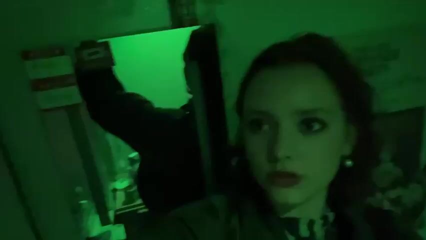 A young woman stands in a room illuminated by green-blue light, taking a selfie. Behind her, reflected in a mirror, is a darker figure of a person wearing a jacket, partially obscured by the dim lighting and reflection. On the wall are nametags and a poster for "The Last Dinner Party." The woman has dark makeup and is wearing a patterned scarf. The scene is cramped and dimly lit.