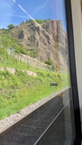The video shows a view from a train window of a rocky hillside with vineyards, as the train travels along the tracks.  A car is visible crossing the train tracks at a railroad crossing.  The sky is blue and sunny.