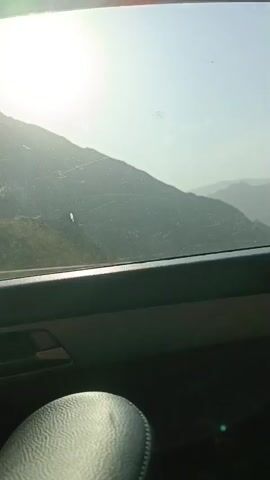 The video appears to be filmed from inside a car, looking out the window at a mountainous landscape. The sun is visible, shining brightly in the sky. The car is moving, as indicated by the slight blur of the scenery. There are some smudges or scratches on the window. The interior of the car is visible in the foreground, with a leather seat and a portion of the dashboard. In the last few frames, a person's head is visible, with a hand holding what appears to be clippers or scissors. The person's head is tilted downwards. It's unclear if the person is cutting their own hair or if someone else is cutting it. The environment outside the car is a natural, undeveloped area with hills and mountains. The lighting suggests it is daytime, possibly late morning or afternoon.