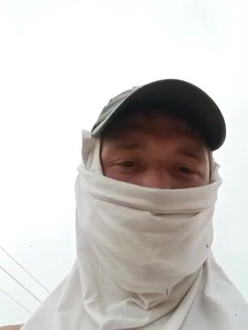 A man wearing a green baseball cap and a white cloth covering the lower half of his face looks into the camera. In the background, two individuals are working on the roof of a brick building. The building appears to be under construction or renovation, with wooden beams visible on the roof. A red tractor or piece of farm equipment is parked near the building. The environment is outdoors, with a cloudy sky and some greenery.