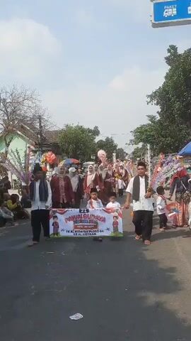 This is a vertical video showing a street parade. The camera is held by someone participating in the parade, looking forward. The parade appears to be a cultural event, possibly involving children from a school, as suggested by a banner held by two adults in the foreground. The banner reads "PAWAI BUDAYA" (Cultural Parade) and has the names "TK AL HIDAYAH PLOSO 04" and "KB AL HISYAA" below it. Two young children in white outfits are walking just behind the banner. Adults in traditional Indonesian clothing, including headscarves and sarongs, are walking in the parade. Some adults are carrying decorative items, possibly symbolic or religious. There are balloons and other festive decorations visible. The parade is taking place on a paved road, with houses and trees lining the sides. Other spectators can be seen on the sidewalks. The weather appears to be sunny. The video is shaky, indicating it was likely recorded while walking.