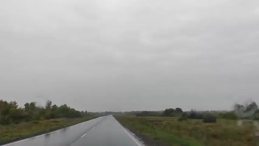 The video shows a view from a car driving down a wet, paved road under a cloudy sky. The road is surrounded by green grass and trees on both sides. The weather appears to be overcast and possibly rainy, as indicated by the wet road and the mood of the sky. There are no people or other vehicles visible in the frame. The perspective suggests the video is captured from the driver's seat or passenger seat of the vehicle. The overall scene depicts a journey through a rural landscape on a dreary day.